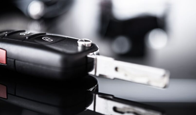 carkeys-autoschluessel-reparatur Ein moderner Autoschlüssel liegt auf einer glänzenden, schwarzen Oberfläche und spiegelt sich darauf. Der Hintergrund ist unscharf und in Schwarz-Weiß gehalten, was dem Bild einen edlen Look verleiht.