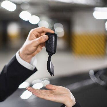carkeys-autoschluessel-reparatur-notfall Ein Mann im Anzug übergibt in einer Tiefgarage einen Autoschlüssel an eine andere Person, deren geöffnete Hand von unten entgegenstreckt. Der Hintergrund ist unscharf und zeigt gelb-schwarze Markierungen an den Wänden.