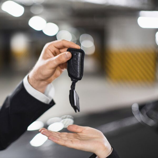 Ein Mann im Anzug übergibt in einer Tiefgarage einen Autoschlüssel an eine andere Person, deren geöffnete Hand von unten entgegenstreckt. Der Hintergrund ist unscharf und zeigt gelb-schwarze Markierungen an den Wänden.