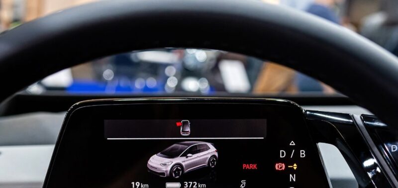 Blick durch das Lenkrad auf das digitale Cockpit eines modernen Elektrofahrzeugs. Auf dem Display wird ein Fahrzeug mit geöffnetem Türsymbol sowie aktuelle Reichweite und Fahrmodus angezeigt.