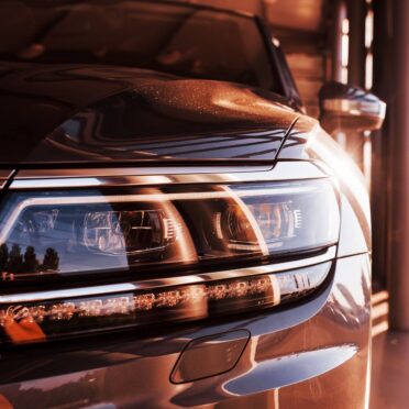 Nahaufnahme des Frontscheinwerfers eines modernen Fahrzeugs mit reflektierendem Metallic-Lack im warmen Licht einer Garage oder Werkstatt.