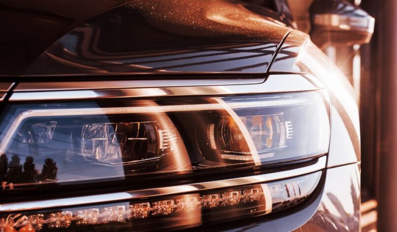 Nahaufnahme des Frontscheinwerfers eines modernen Fahrzeugs mit reflektierendem Metallic-Lack im warmen Licht einer Garage oder Werkstatt.