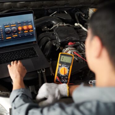 Ein Kfz-Techniker überprüft den Motorraum eines Fahrzeugs mithilfe eines Laptops mit Diagnosesoftware und eines digitalen Multimeters. Der Fokus liegt auf der technischen Analyse und elektronischen Fehlerdiagnose.