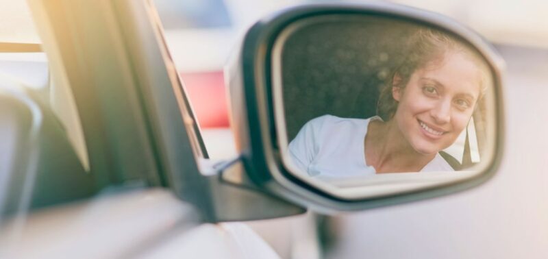 Eine Frau lächelt aus dem Autoinneren und ist deutlich im Seitenspiegel eines weißen Fahrzeugs zu sehen. Die Szene ist von weichem Sonnenlicht durchflutet und vermittelt eine positive, entspannte Stimmung.