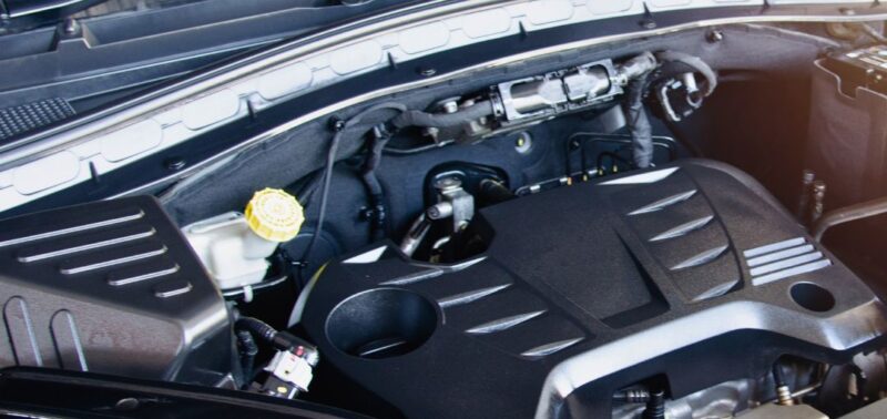 Blick in den geöffneten Motorraum eines Fahrzeugs mit modernem Motorblock. Verschiedene Bauteile wie die Abdeckung, Verkabelung und Flüssigkeitsbehälter sind deutlich sichtbar.