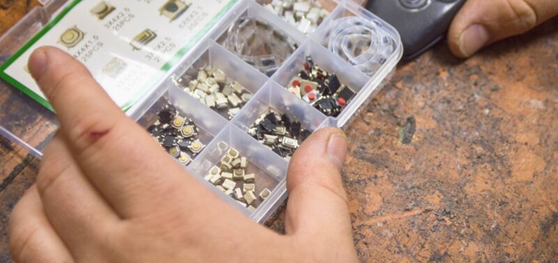 Zwei Hände auf einer Werkbank – eine hält ein Autoschlüssel-Gehäuse, die andere eine Sortierbox mit verschiedenen Mikrotastern und elektronischen Kleinteilen. Die Szene deutet auf eine Reparaturarbeit am Schlüssel oder Zündsystem hin.